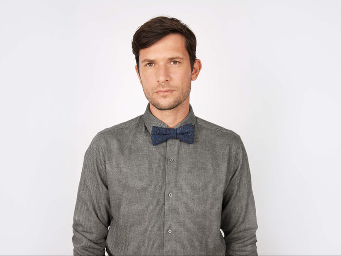 A man with short brown hair wears a gray button-up shirt and the Hanna Hats Bow Tie Tweed, standing against a plain white background and looking directly at the camera with a neutral expression.