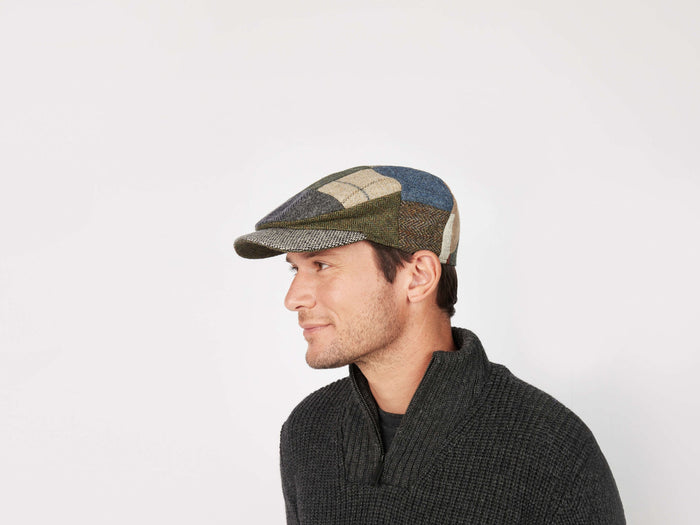 A man in a dark textured sweater and a multicolored Daithi Cap Patchwork Tweed by Hanna Hats looks left against a plain white background, highlighting the vibrant style of this Irish tweed cap.