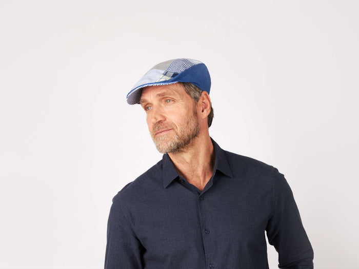 A middle-aged man with a beard wears a Hanna Hats Donegal Touring Cap Patchwork Grey/Blue Linen and a dark blue button-up shirt, looking thoughtfully to the side against a plain white background.