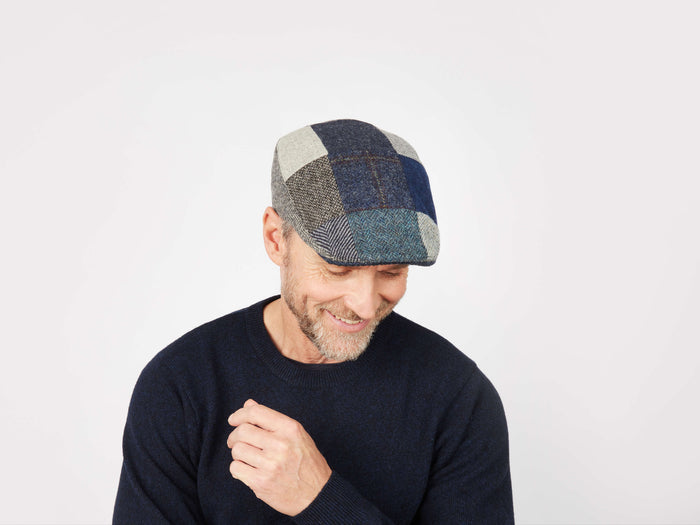 A man wearing a dark sweater and the Hanna Hats Donegal Touring Cap Patchwork Grey/Blue Tweed smiles while looking down against a plain white background.