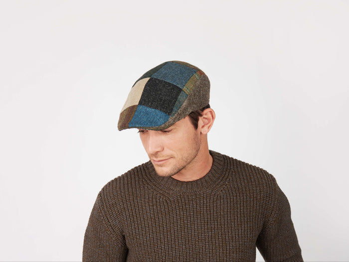 A man in a brown textured sweater and a Hanna Hats Donegal Touring Cap Patchwork Tweed looks down and slightly to his left against a plain white background.