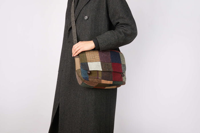 A person in a long, dark herringbone coat holds the Hanna Hats Satchel Bag Patchwork Tweed against a plain light background.