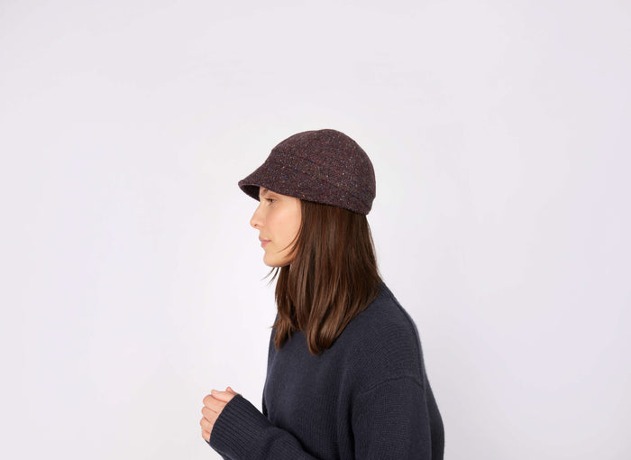 A woman with straight brown hair in a dark knit sweater and a Hanna Hats Slieve League Hat Tweed stands in profile against a plain white background.