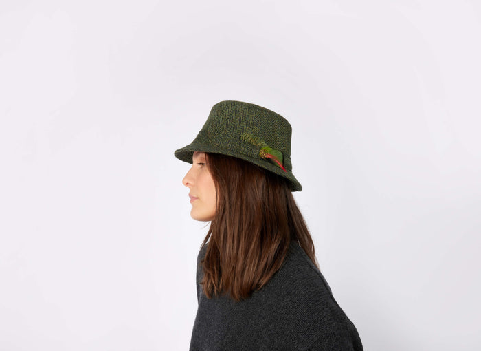 A person with straight brown hair, seen in profile against a white background, wears the Hanna Hats Walking Hat Tweed—a green Irish hat with a feather, crafted from authentic Irish tweed.